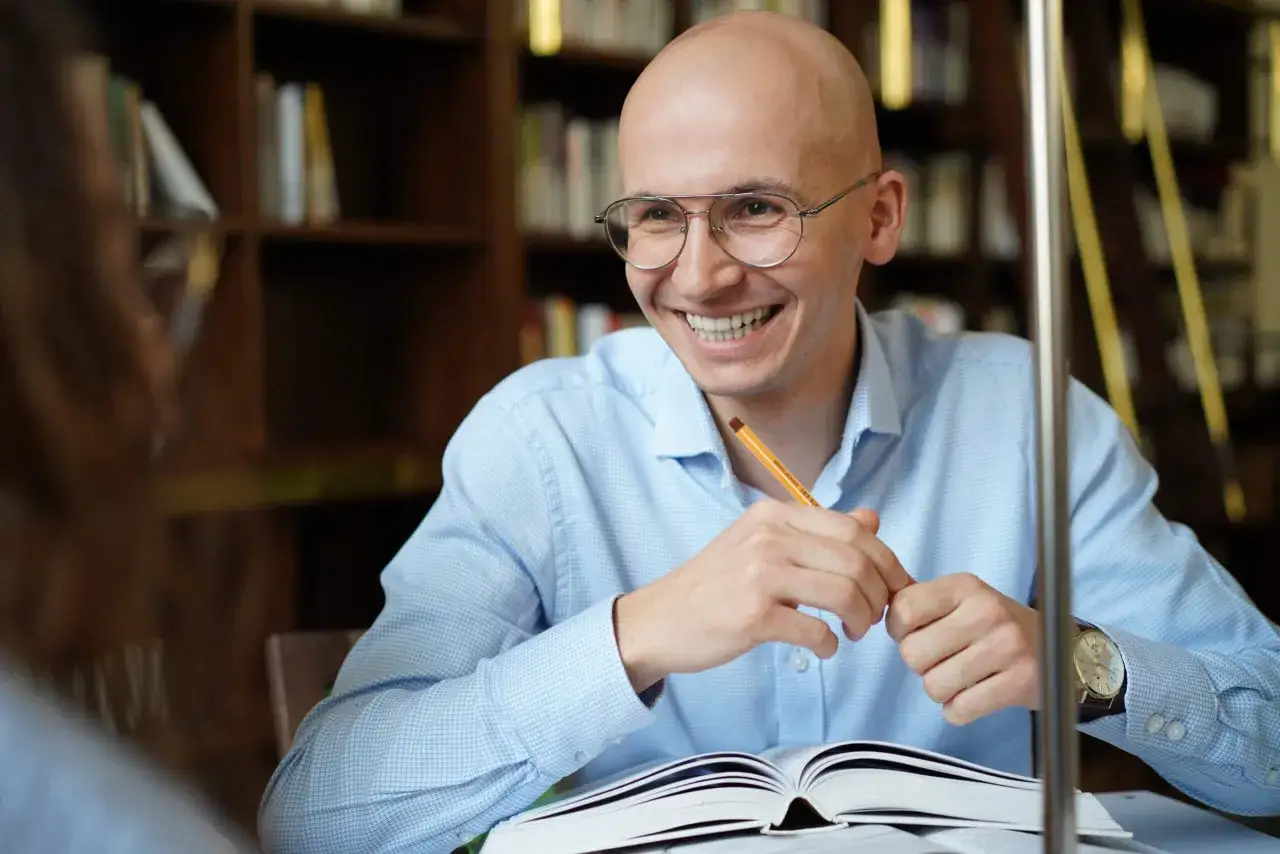 Uśmiechnięty doktorant w okularach, z oł&oacute;wkiem w dłoni, pochyla się nad otwartą księgą. Tak wyglądają studia doktoranckie &ndash; pełne pasji i nauki.