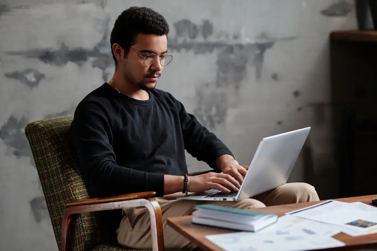 En ung man med glas&ouml;gon sitter i en f&aring;t&ouml;lj och arbetar p&aring; sin laptop. Han verkar vara en frilansare som njuter av flexibiliteten i sitt arbete.