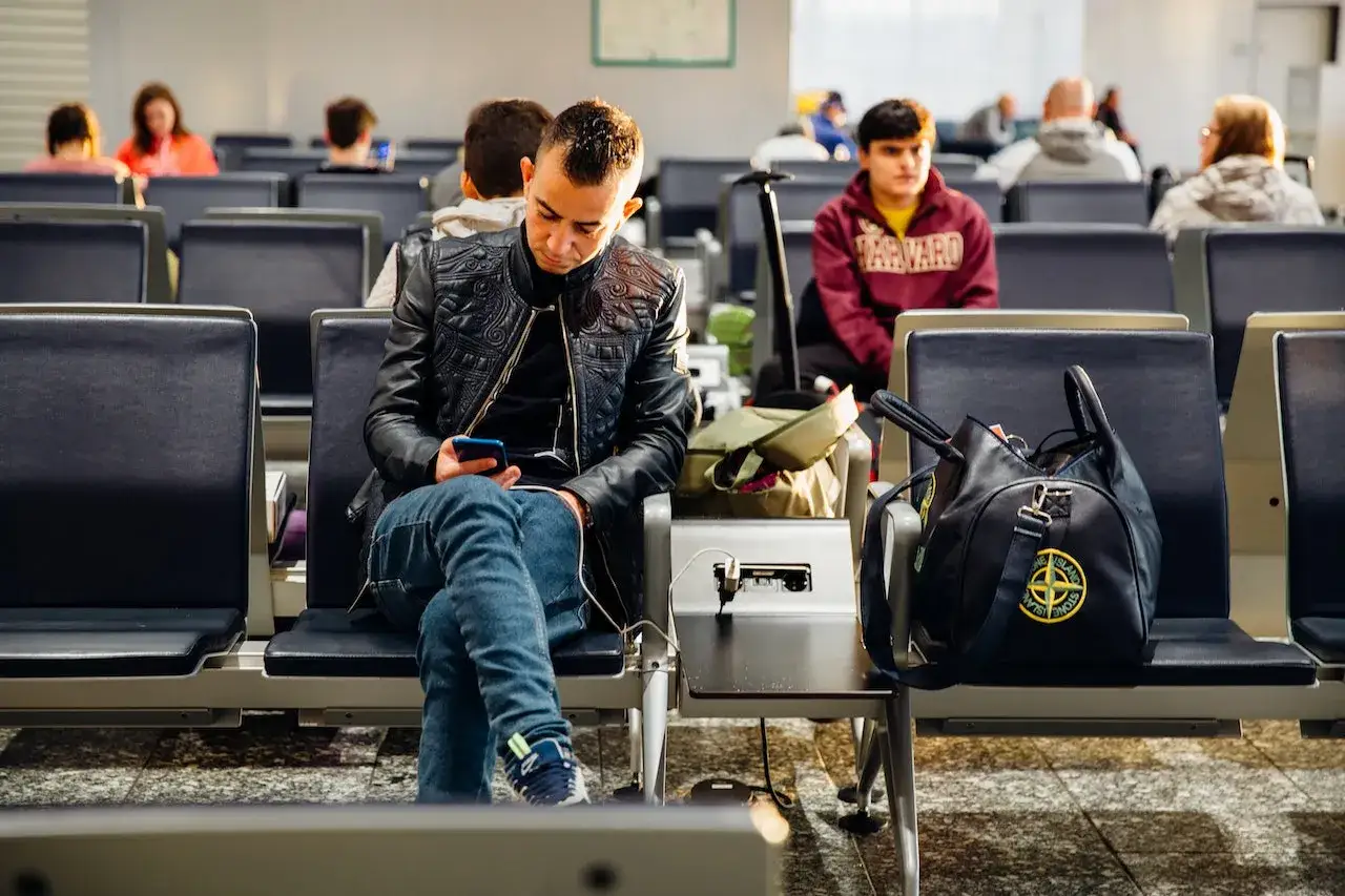 Mężczyzna w czarnej kurtce siedzi na lotnisku, sprawdzając telefon. Zastanawia się, odprawa online ile wcześniej na lotnisku jest najlepsza.