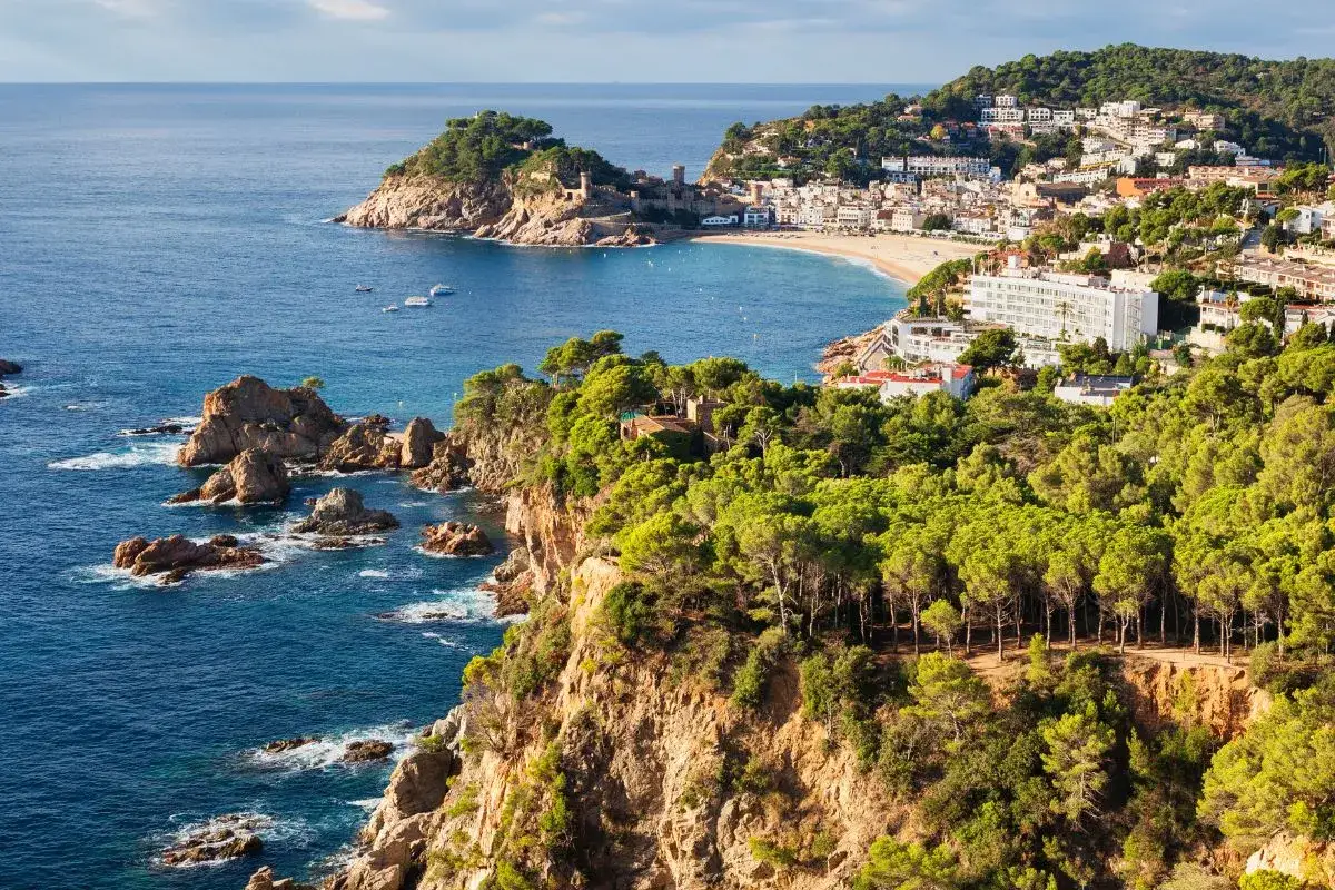 Coastal town with a beach and castle, perfect for a road trip from Barcelona. Lush green cliffs meet the blue sea.