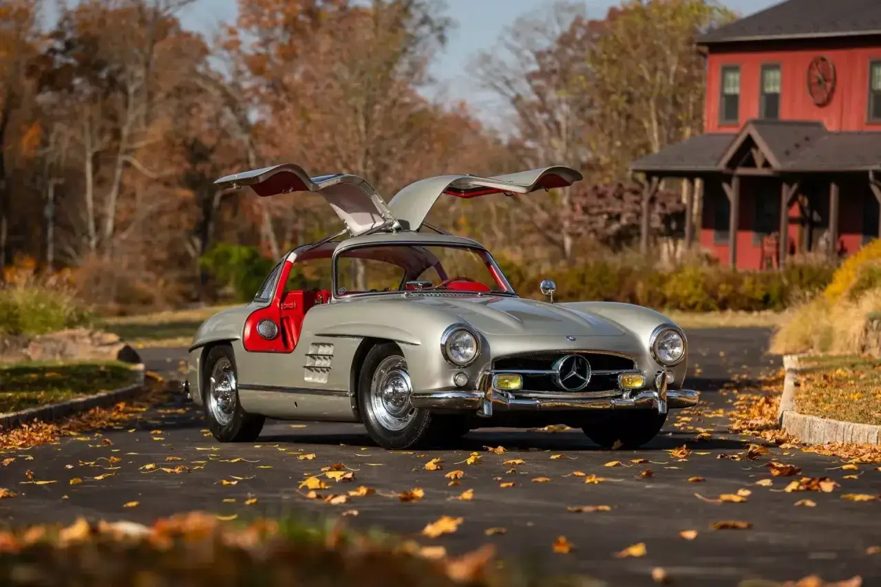Srebrny Mercedes 300 SL z otwieranymi do góry drzwiami, na tle jesiennego krajobrazu i czerwonego domu.