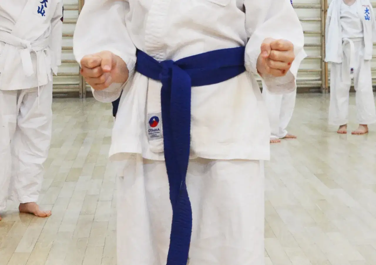 Dziecko w białym karate gi z niebieskim pasem, symbolizującym stopień mistrzowski w karate, gotowe do treningu.