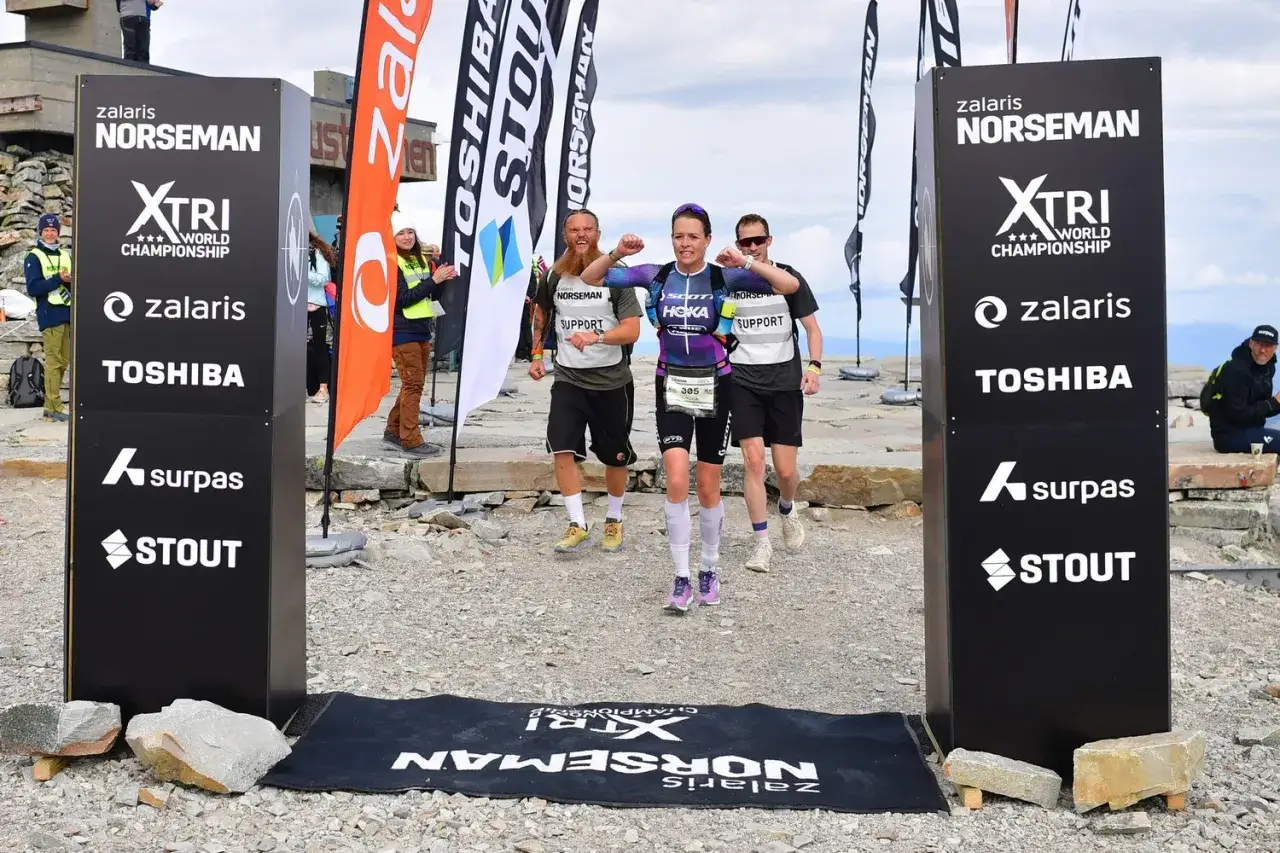 L&auml;uferinnen und L&auml;ufer erreichen das Ziel beim Norseman Triathlon. Die Siegerin jubelt, gefolgt von zwei m&auml;nnlichen Teilnehmern.