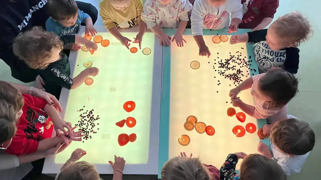 Bambini esplorano fette di agrumi e semi su tavoli luminosi, un'attività che stimola la costruzione del sé nel bambino attraverso il gioco sensoriale.