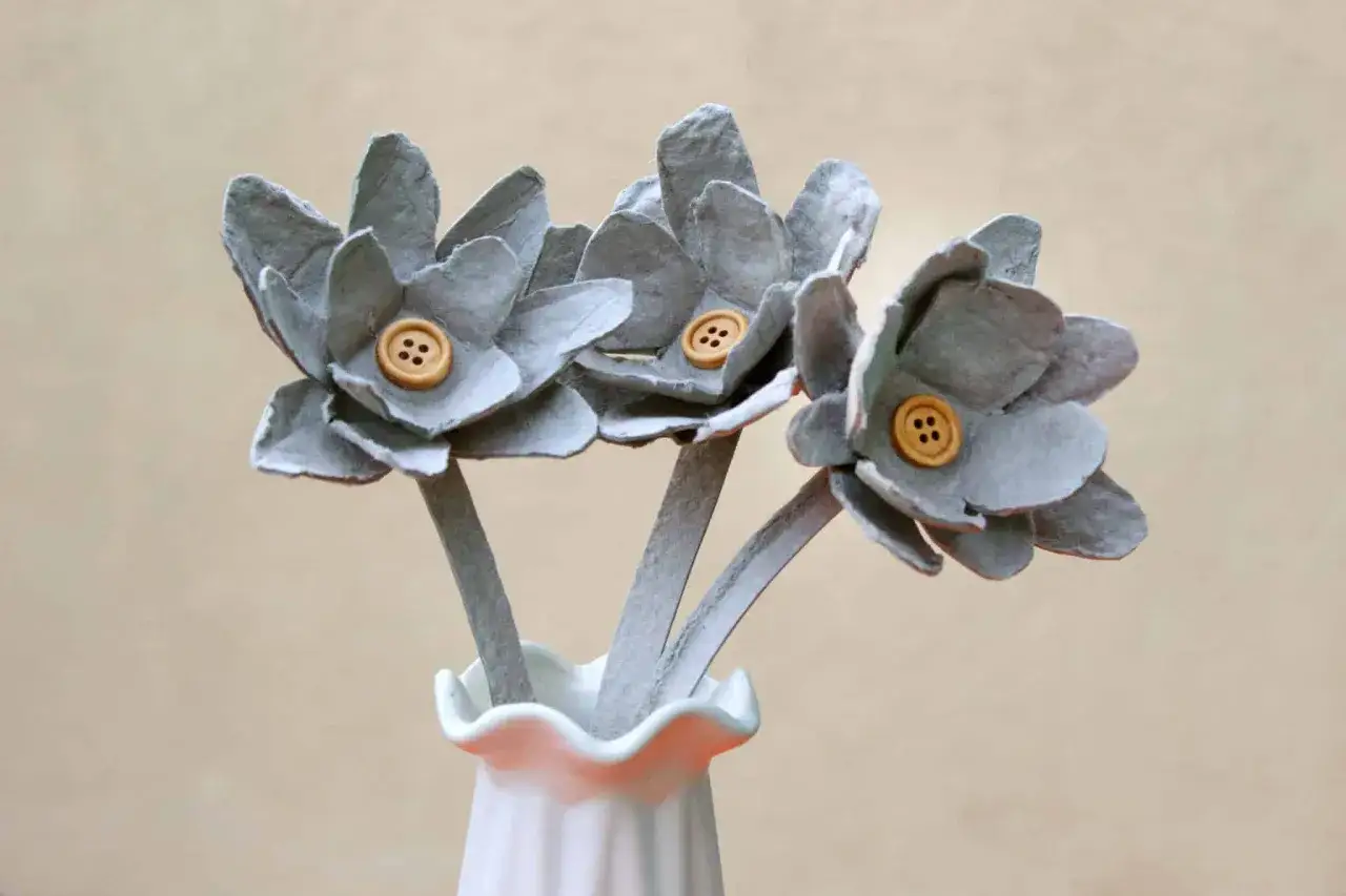 Graue Blumen aus Eierkarton mit Holzkn&ouml;pfen als Mitte stehen in einer wei&szlig;en Vase.