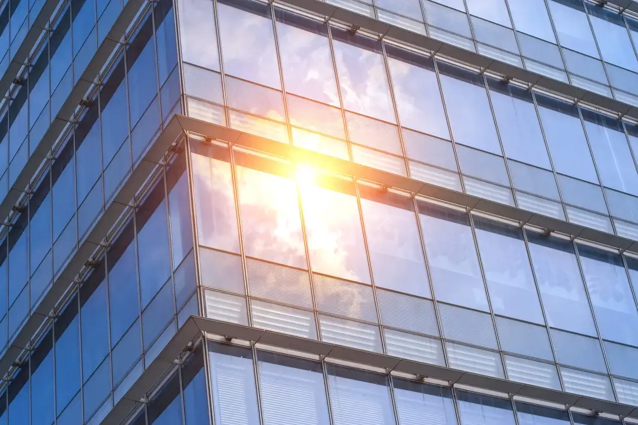 Edificio moderno con fachadas de **solar glass** que reflejan el cielo azul y el sol brillante.