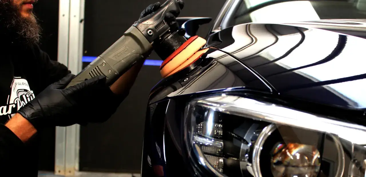 A detailer uses a polisher to cut and buff a dark car's fender, bringing out a mirror-like shine.
