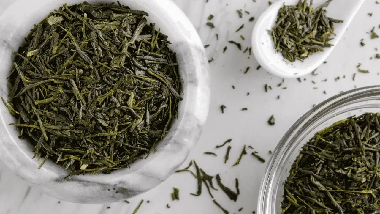 Trockene Sencha-Teebl&auml;tter in einer Marmorschale und einem L&ouml;ffel. Die **Sencha Tee Wirkung** ist belebend und erfrischend.