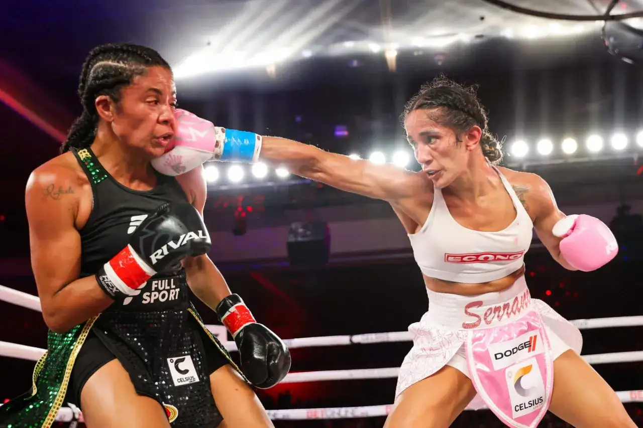 Two professional female boxers exchange punches in a brightly lit ring. One boxer, wearing black and green, is hit on the cheek.