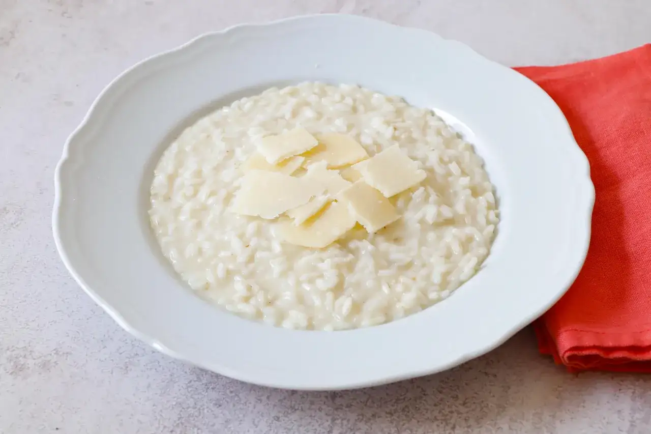 Un cremoso riso in bianco, condito con scaglie di parmigiano, servito in un piatto bianco su un tovagliolo rosso.