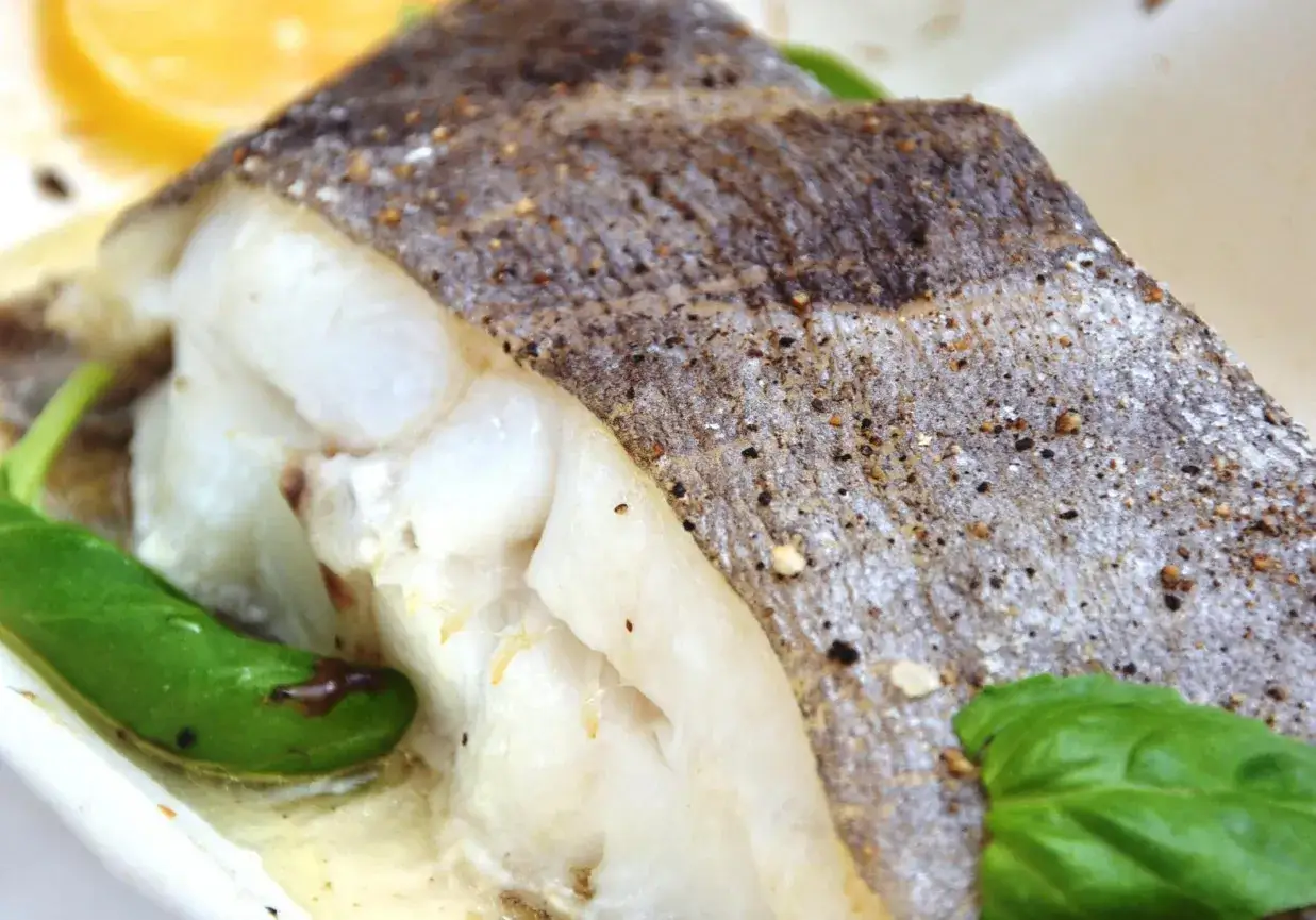Soczysty halibut pieczony ze skórą, doprawiony pieprzem i podany z listkami bazylii i plasterkiem cytryny.