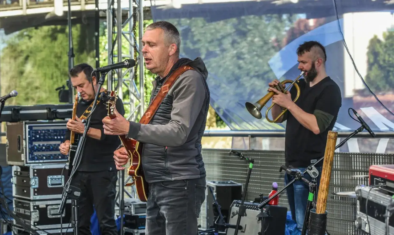 Wokalista zespołu Raz Dwa Trzy śpiewa, grając na gitarze. Obok niego gitarzysta i trębacz.