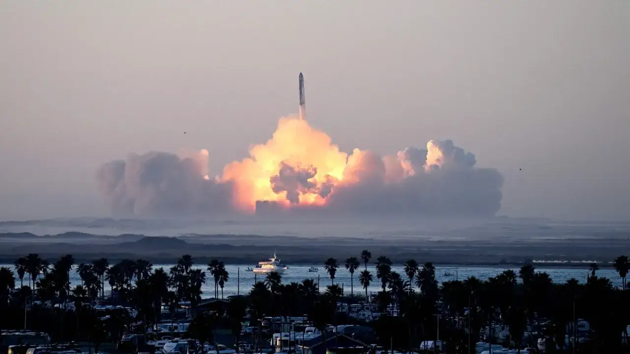 Starship start w chmurach ognia i dymu, nad palmami i wodą. Statek kosmiczny wznosi się w niebo.