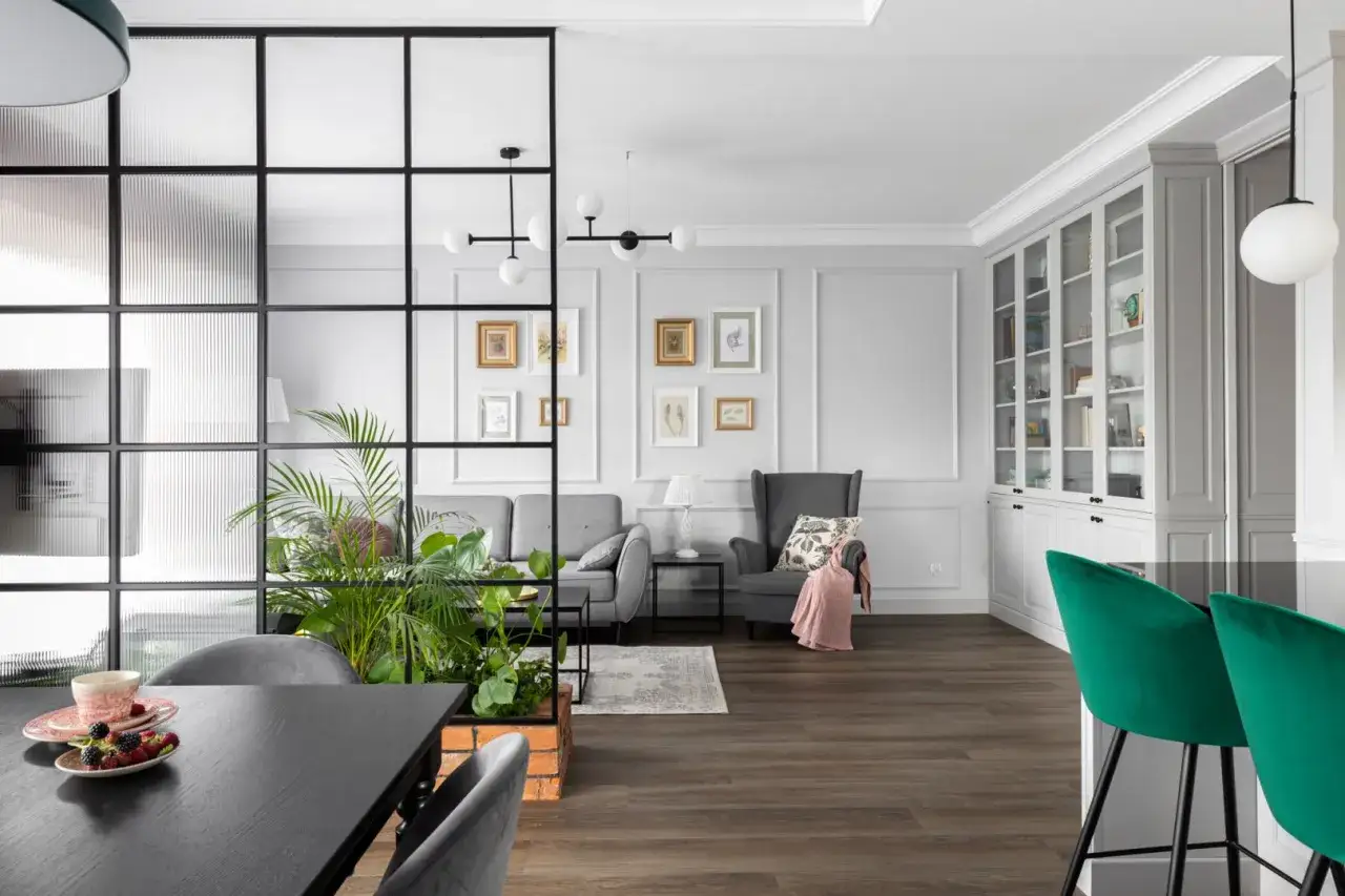 Wnętrze z przeszkloną ścianką działową, za którą widać bujny kwiat w doniczce. W salonie szara sofa, fotel i regał.