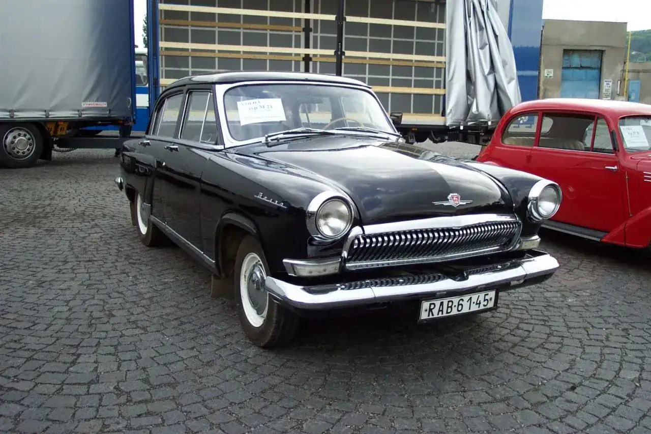 Svart Volga GAZ-21, en klassisk sovjetisk bil, st&aring;r parkerad p&aring; kullerstensbel&auml;ggning. En r&ouml;d bil syns i bakgrunden.