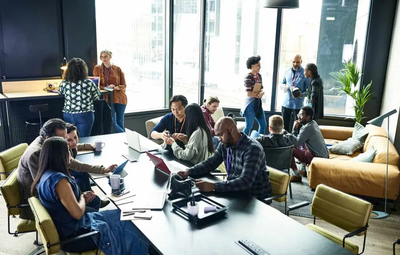 Un formateur web anime une session de travail collaborative dans un espace moderne. Des participants &eacute;changent autour d'une table, certains sur leurs ordinateurs.