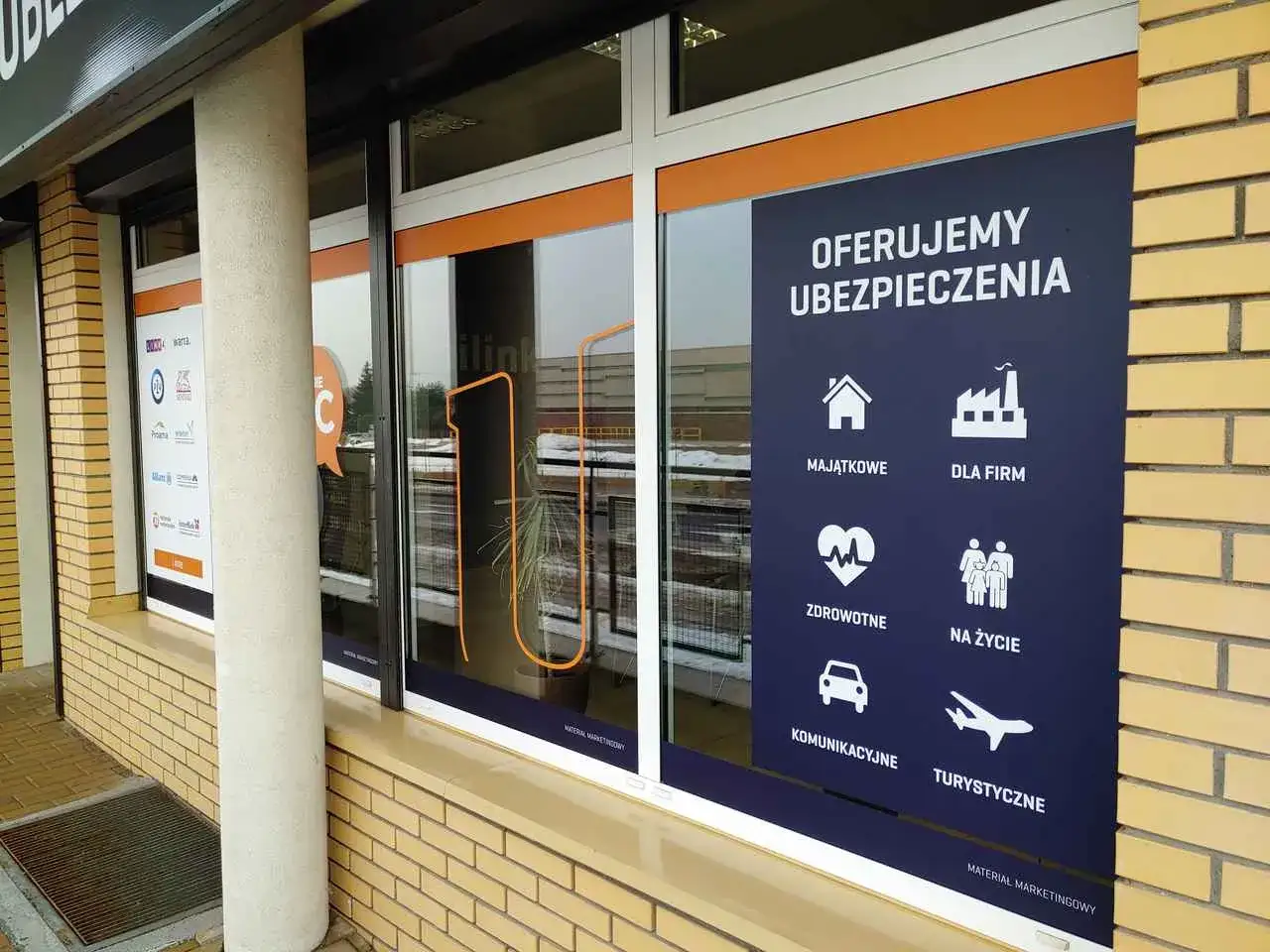 Plakat na witrynie sklepowej z ofertą ubezpieczeń: majątkowe, dla firm, zdrowotne, na życie, komunikacyjne, turystyczne.