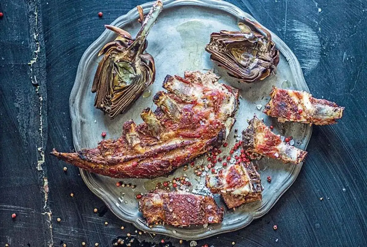 Un plateau de côtes d'agneau grillées et deux moitiés d'artichaut au barbecue, saupoudrés de poivre rose et de gros sel.