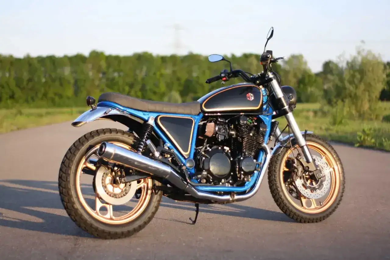 Niebieski motocykl Yamaha XJ 900 w stylu cafe racer, z miedzianymi felgami i chromowanym wydechem, stoi na drodze.