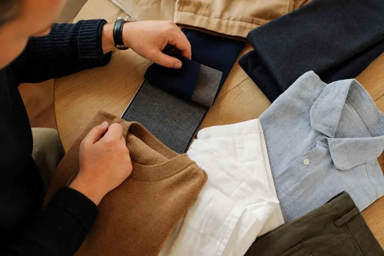 Mann w&auml;hlt Stoffe f&uuml;r seine Capsule Wardrobe Men aus: brauner Pullover, wei&szlig;e und blaue Hemden, dunkle Hose und Stoffmuster.