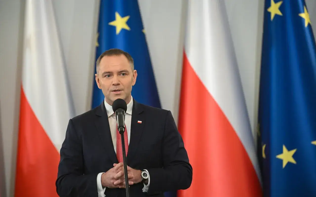 Mężczyzna w garniturze przemawia na tle flag Polski i UE. Czy to wyjaśnia, dlaczego prezydent nie skorzystał z prawa łaski wobec skazanych urzędników państwowych?