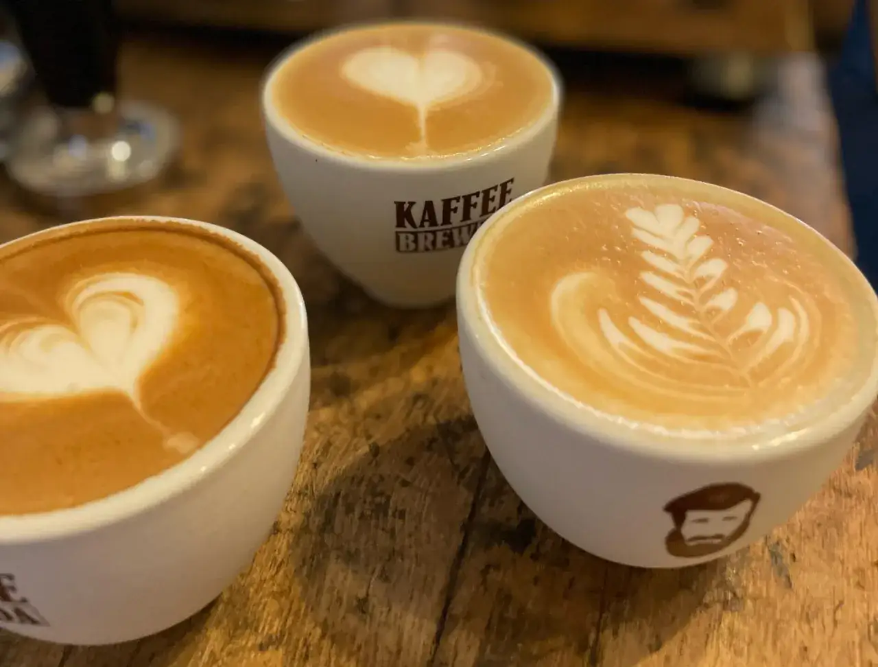 Drei Kaffees mit Latte Art: Ein Herz, ein Blatt und ein Flat White vs. Cappuccino Vergleich auf Holztisch.