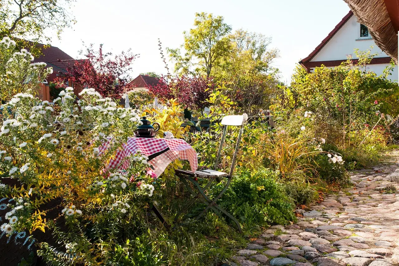Ein Tisch mit karierter Decke und Teekanne l&auml;dt im naturnahen Garten zum Verweilen ein. &Uuml;ppige Blumen und B&auml;ume umgeben den Sitzplatz.