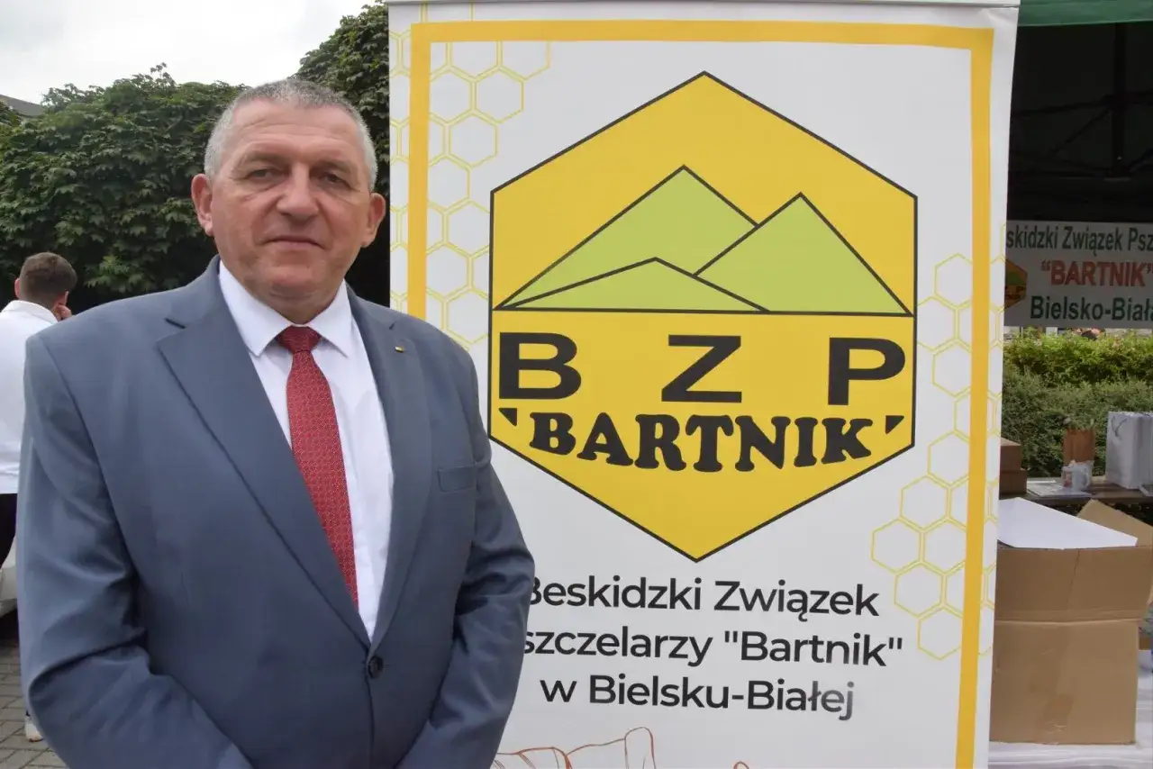 Elegancki mężczyzna w garniturze, dawny pszczelarz, stoi obok banera Beskidzkiego Związku Pszczelarzy