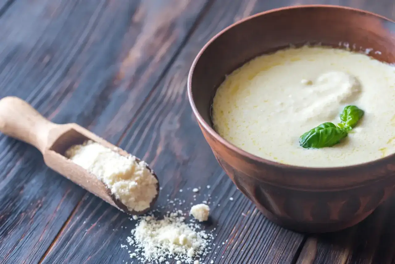 Bol en terre cuite rempli d'une onctueuse sauce parmesan italienne, garnie de basilic frais. Une cuill&egrave;re en bois contient du parmesan r&acirc;p&eacute;.