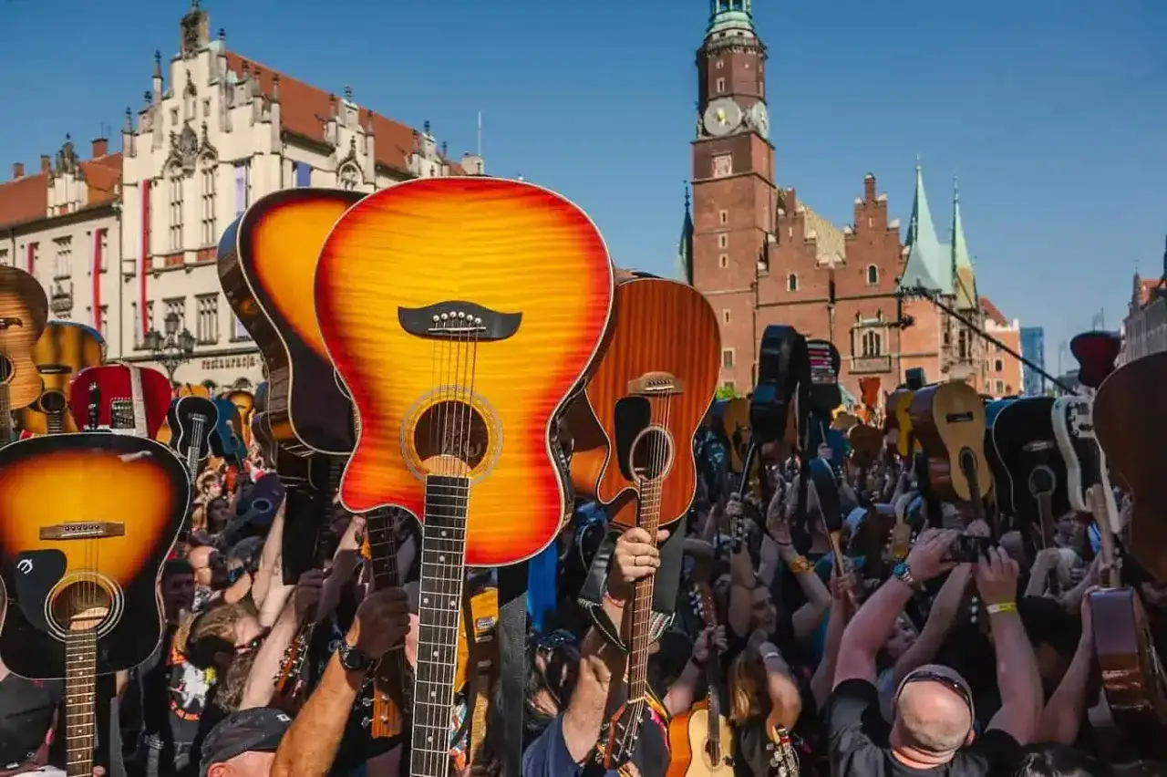 Tłum ludzi z gitarami wzniesionymi ku g&oacute;rze, świętujący gitarowy rekord świata. W tle zabytkowa zabudowa miejska.