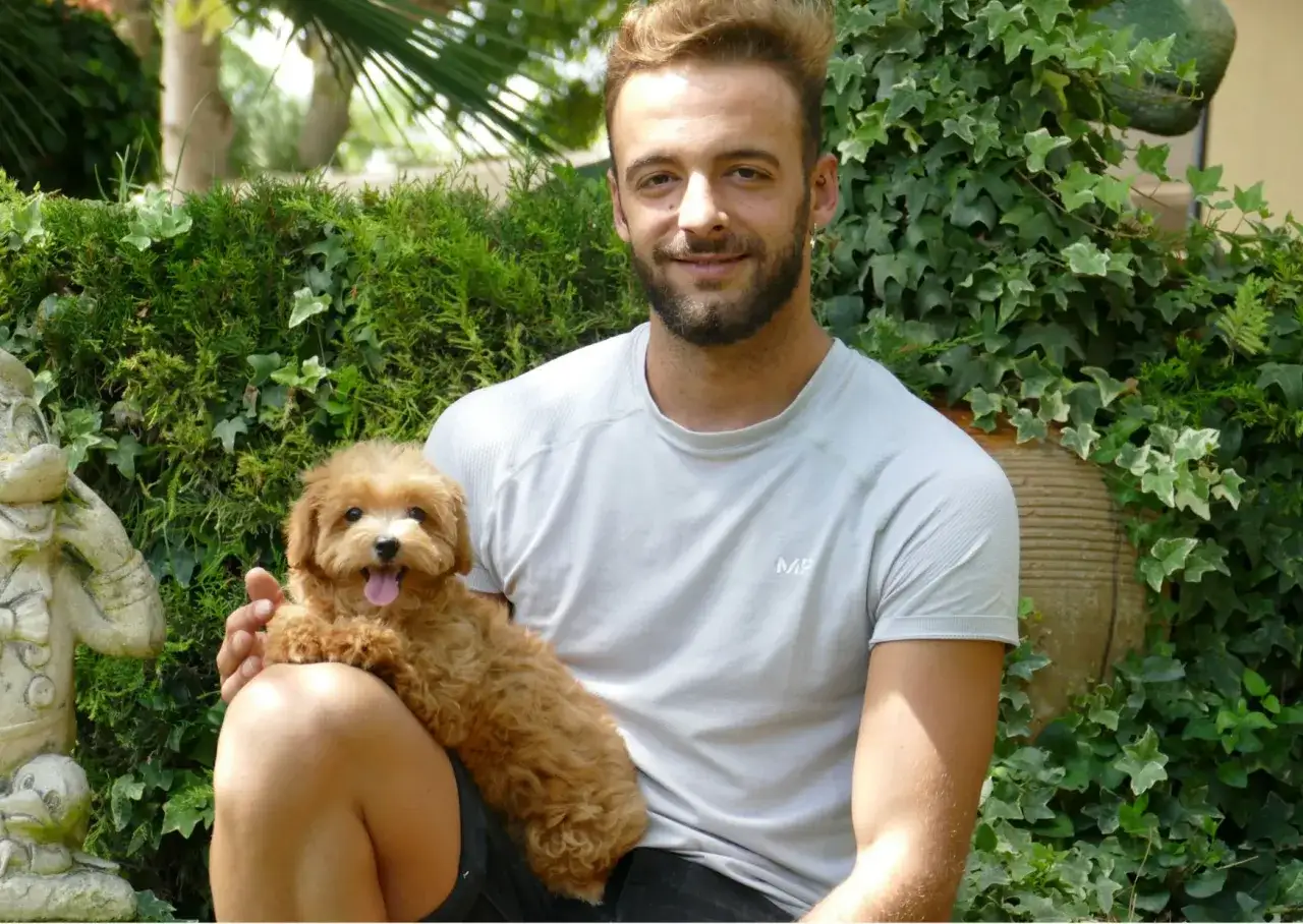 Hombre sonriente abraza a un maltipoo adulto tama&ntilde;o, con pelaje rizado y color caramelo, en un jard&iacute;n exuberante.