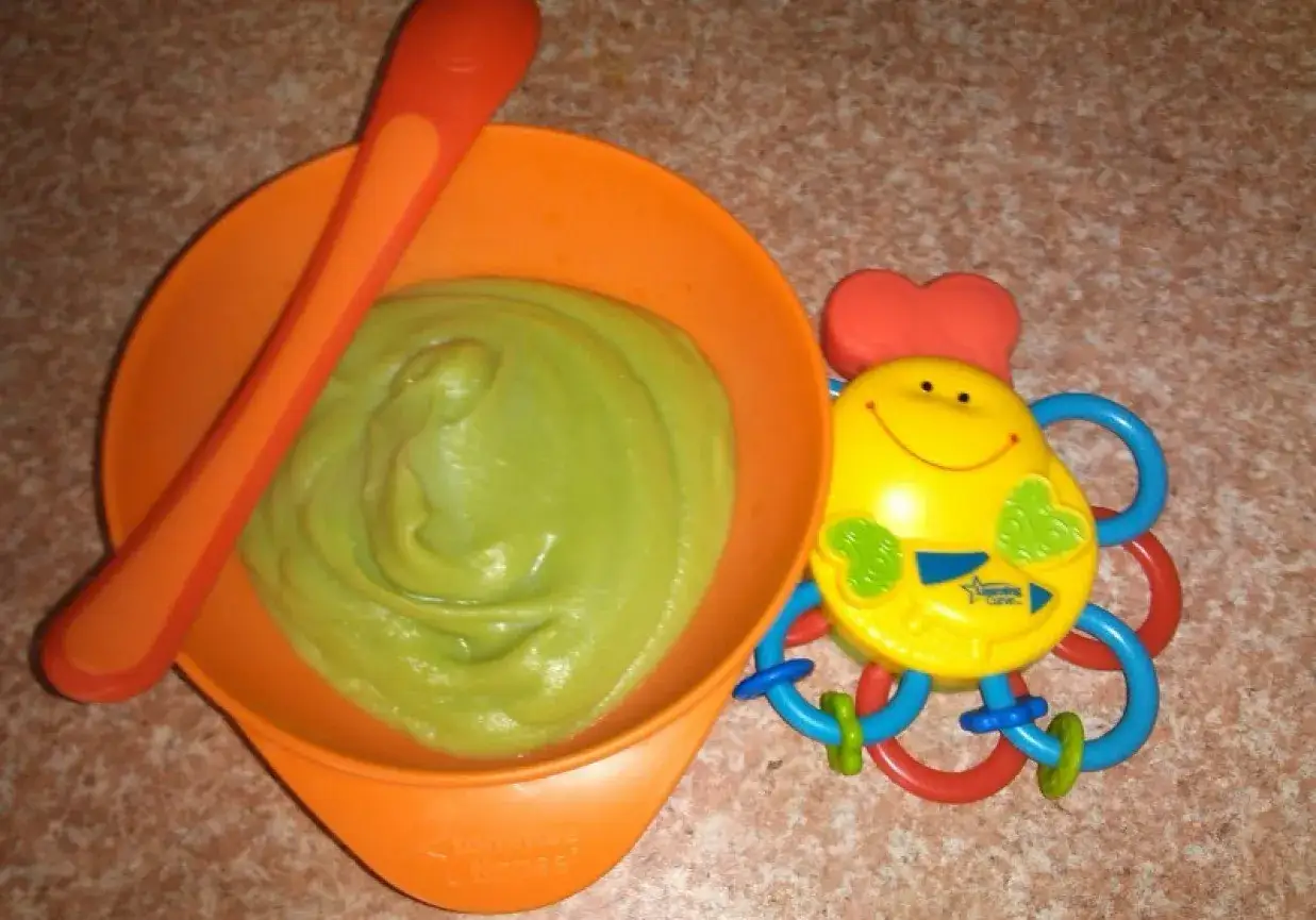 Miseczka z zielonym puree, idealna odpowiedź na pytanie z czym łączyć brokuł dla niemowlaka. Obok zabawka-pajacyk.
