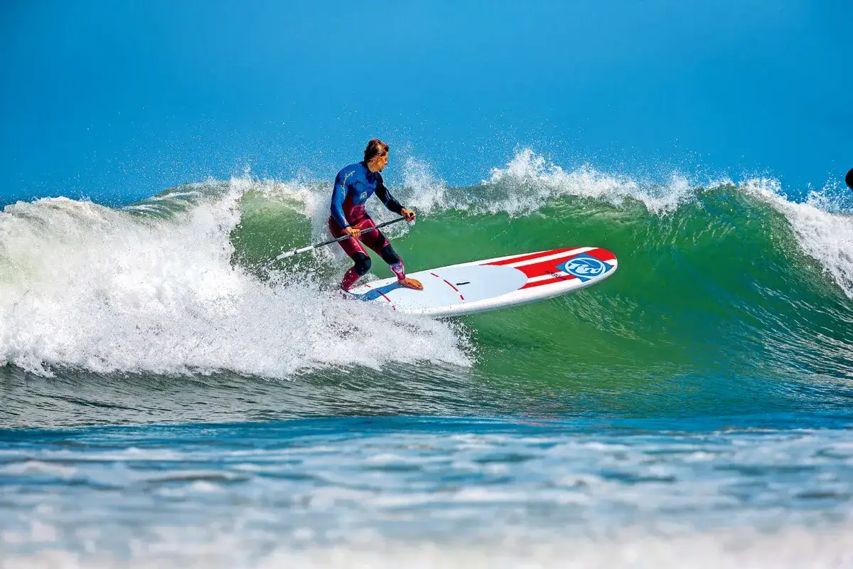 Ein Mann im Neoprenanzug surft auf einem SUP-Board eine grüne Welle. Das SUP-Wellenreiten erfordert Balance und Geschick.