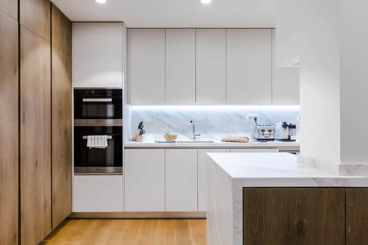 Cocina moderna con gabinetes blancos y madera. La iluminación LED bajo los gabinetes superiores resalta el salpicadero de mármol, mostrando cómo iluminar una cocina con estilo.
