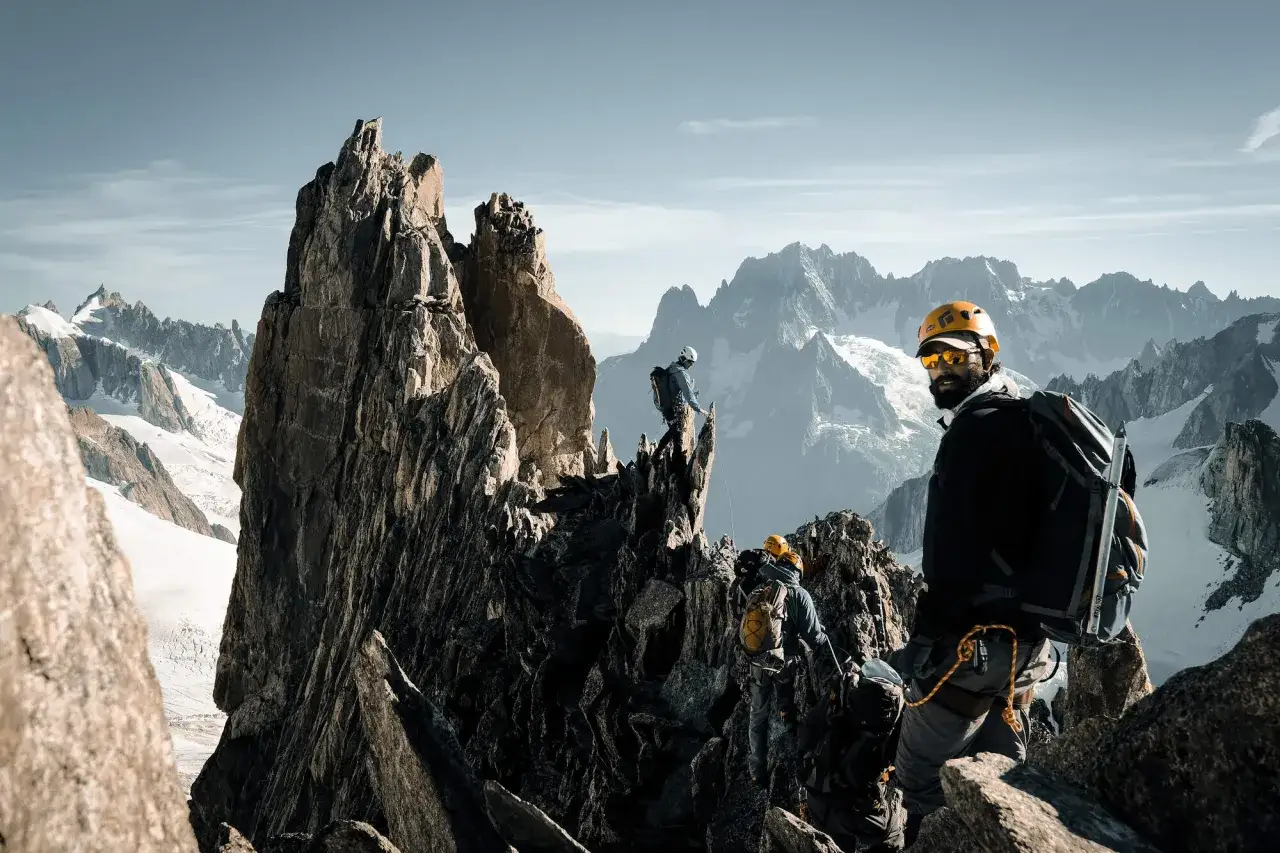 Antoine Cayrol et son équipe escaladent un pic rocheux escarpé, avec des montagnes enneigées en arrière-plan.
