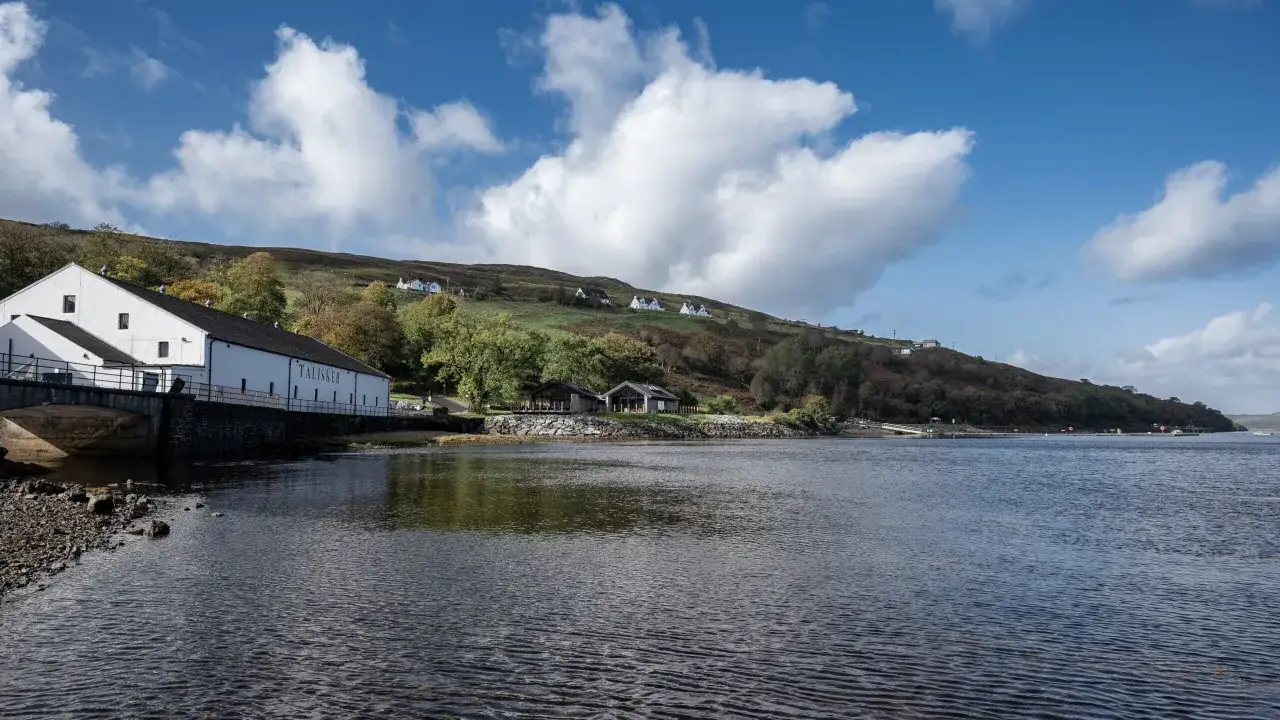 La distillerie Talisker, avec ses bâtiments blancs, se dresse au bord d'une baie écossaise sous un ciel bleu et nuageux.
