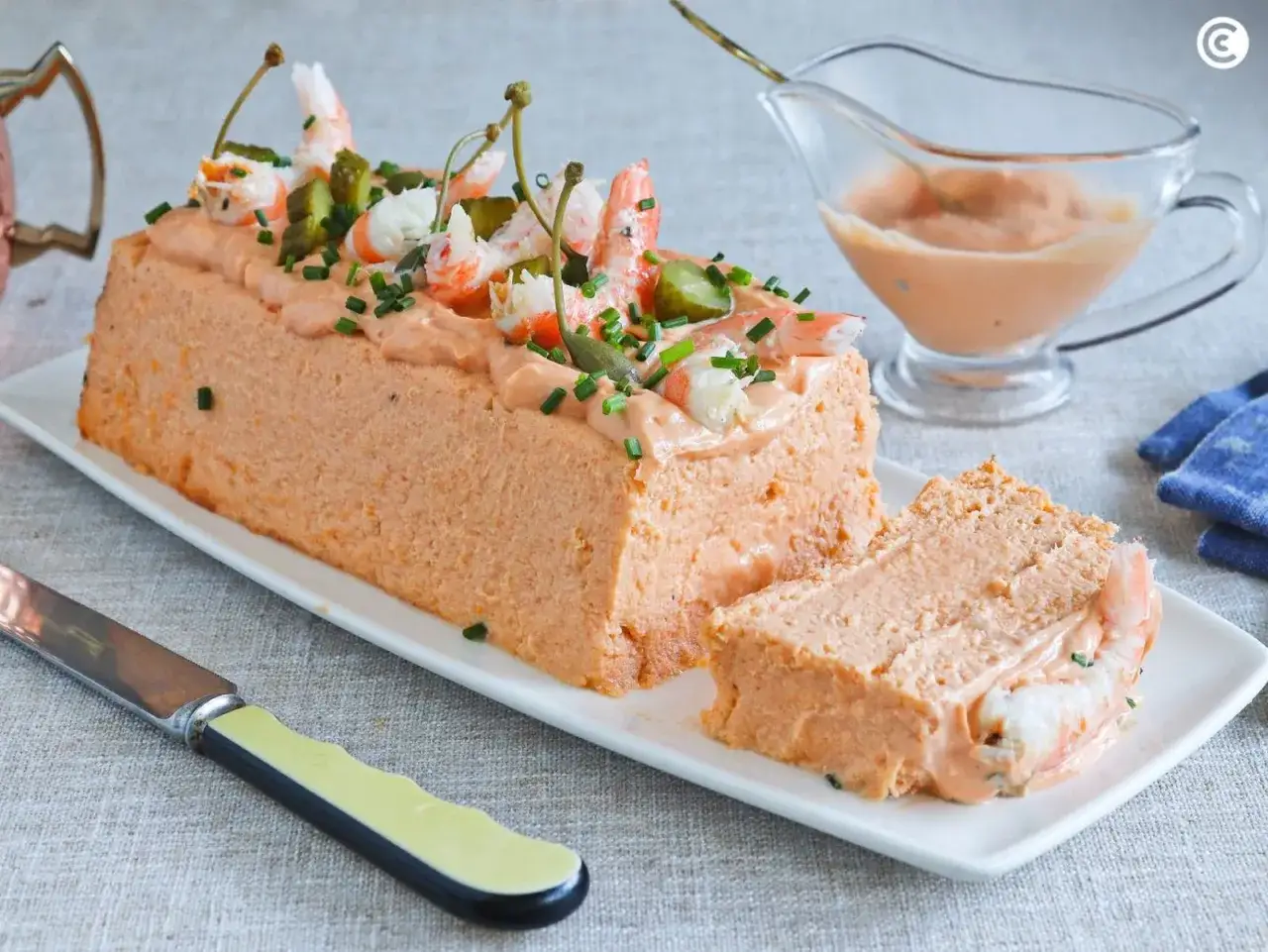 Pastel de pescado decorado con gambas, pepinillos y alcaparras. Una porci&oacute;n ya cortada muestra el interior cremoso.