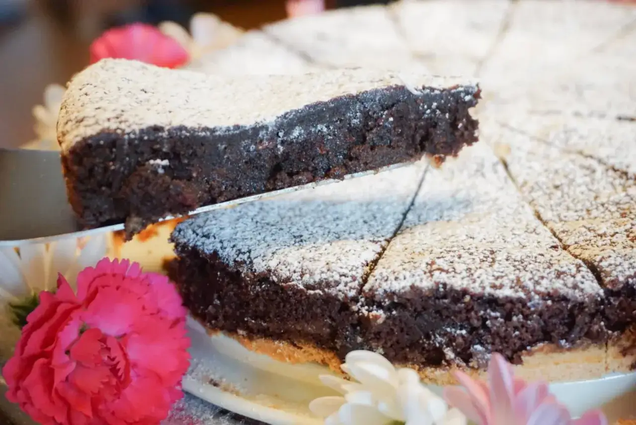 En bit av en härlig glutenfri kladdkaka lyfts upp. Den är pudrad med florsocker och dekorerad med rosa och vita blommor.