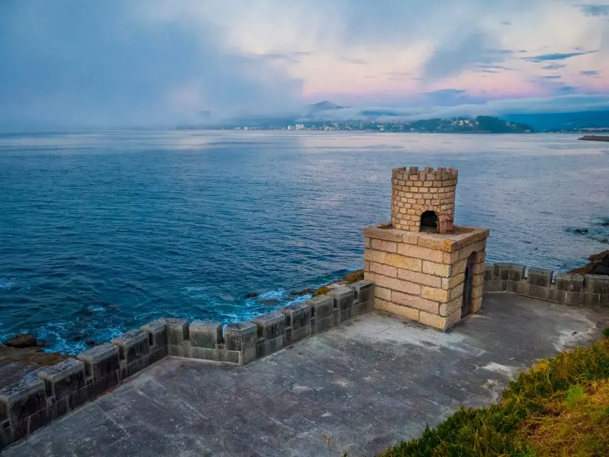 Torreón de piedra junto al mar, con vistas a pueblos cerca de Malpica y un cielo nublado al amanecer.