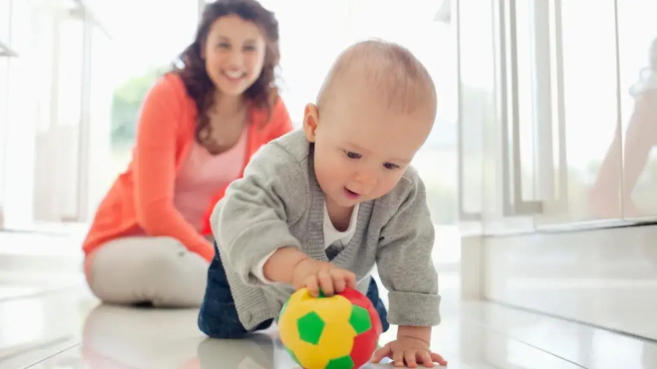 Bebé gateando hacia una pelota de colores, disfrutando del movimiento libre mientras su madre sonríe al fondo.
