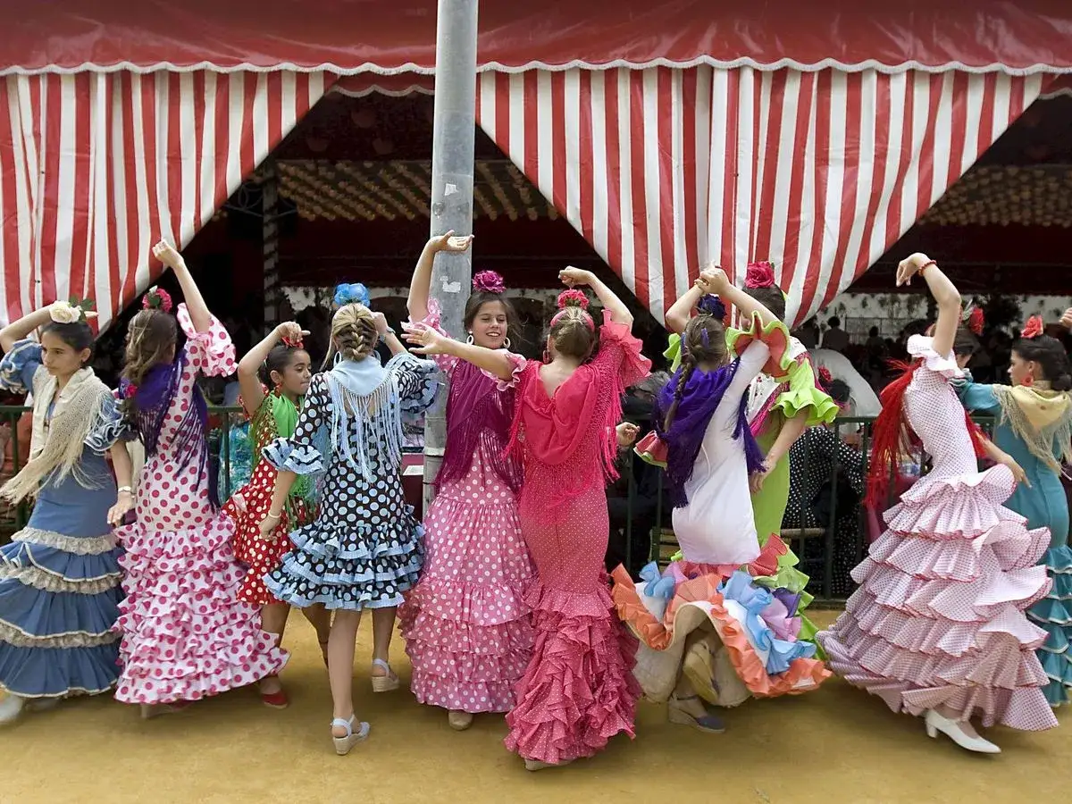 Ni&ntilde;as con trajes de lunares y volantes, listas para bailar sevillanas bajo una carpa a rayas.