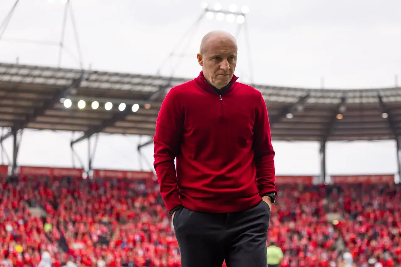 Nowy trener Widzewa w czerwonym swetrze idzie po murawie, w tle trybuny wypełnione kibicami.