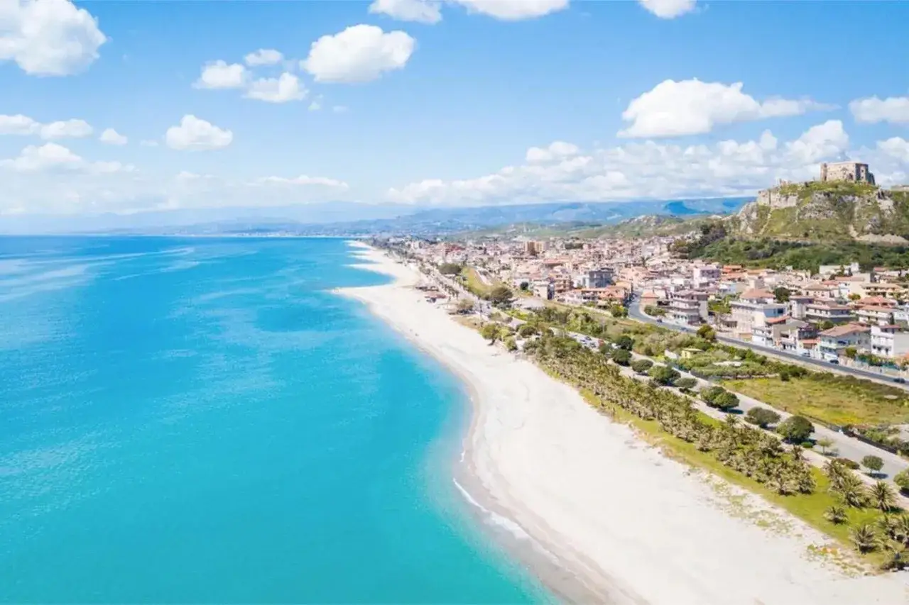 Spiaggia bianca e mare turchese con il Valtur Otium Resort sullo sfondo, un borgo con castello arroccato su una collina.