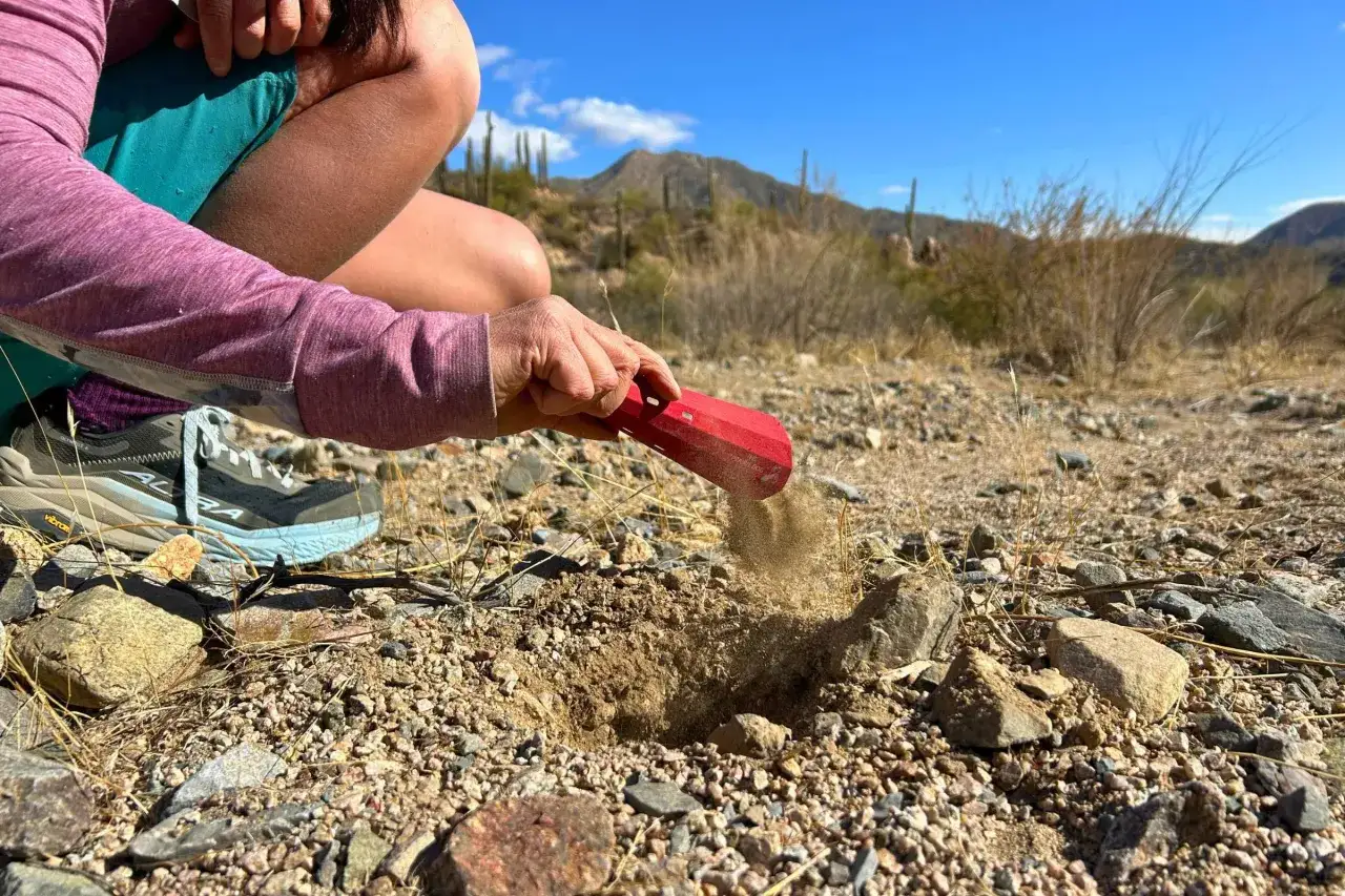 A hiker uses a red trowel to bury trash in the desert, leaving no trace behind.