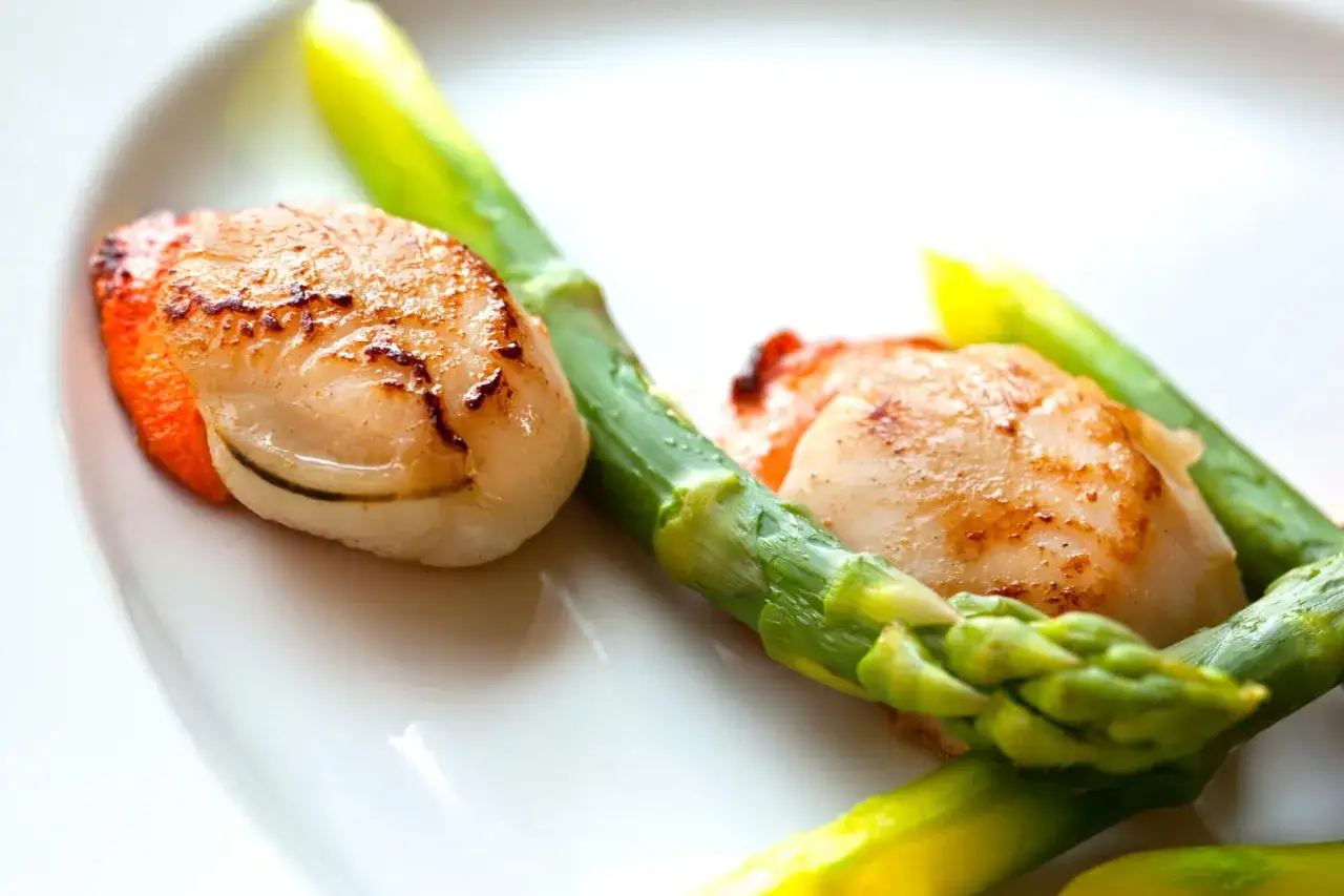 Deux noix de Saint-Jacques poêlées, tendres et dorées, accompagnées de pointes d'asperges vertes croquantes sur une assiette blanche.