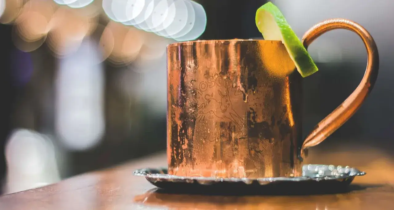 Miedziany kubek z Moscow Mule, ozdobiony plasterkiem limonki, stoi na metalowej podstawce. Obok, w tle, majaczy London Mule.