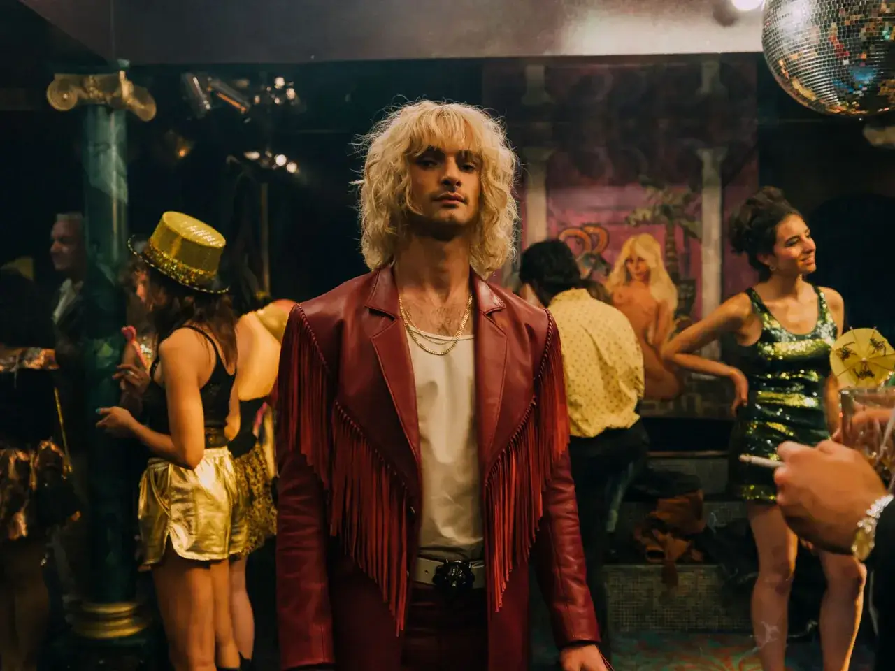 Ein Mann mit blonden Haaren und einer roten Fransenjacke steht im Vordergrund. Im Hintergrund sind tanzende Menschen und eine Discokugel zu sehen. Die Szene erinnert an die Besetzung von Luden.