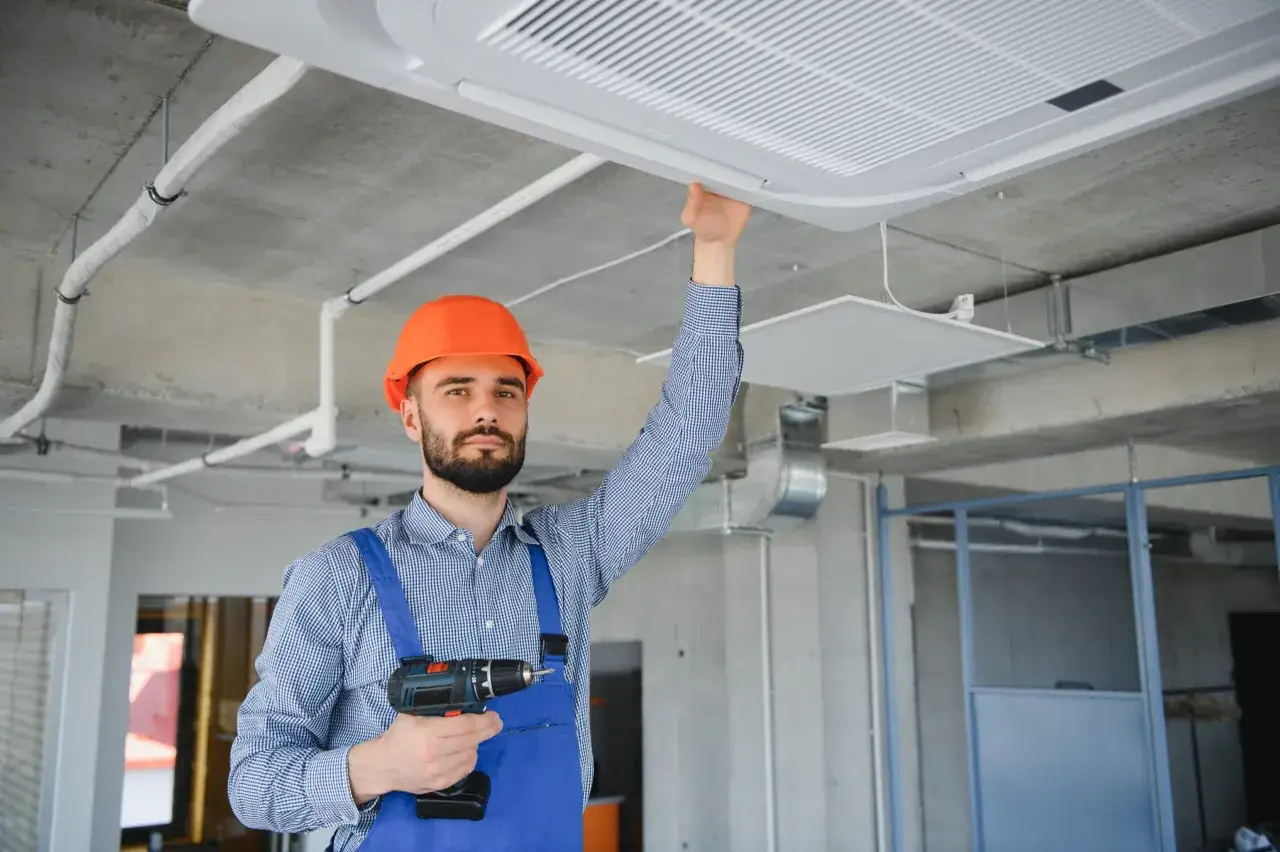Instalator w kasku i kombinezonie montuje klimatyzator pod sufitem, trzymając wiertarkę.