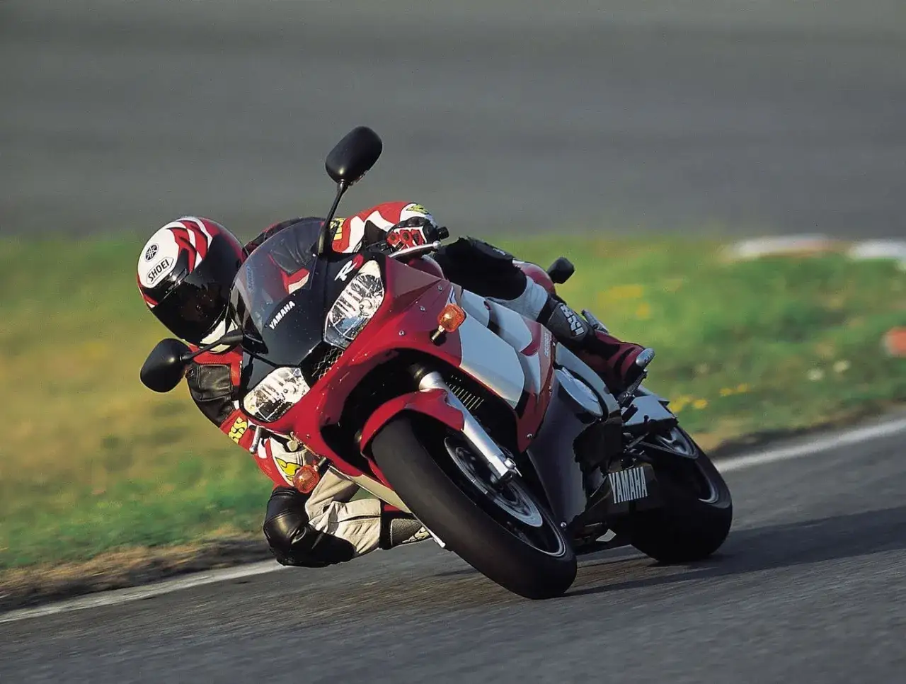 Motocyklista na torze wyścigowym pochyla się na swoim czerwono-białym motocyklu Yamaha R6 2003, pokonując zakręt z dużą prędkością.