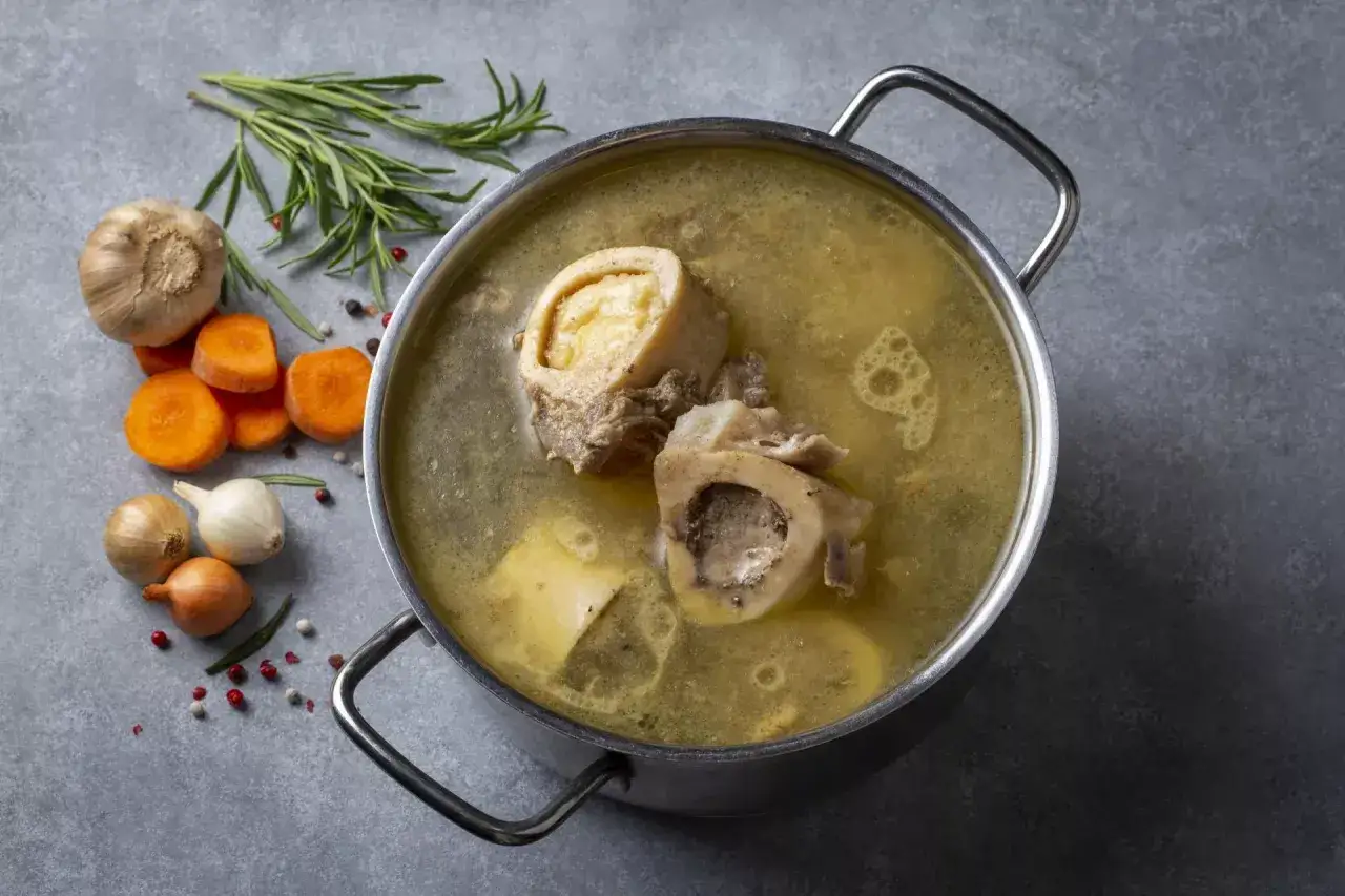 Aromaticzny wywar z kości szpikowych w garnku, obok marchewka, cebula, czosnek i rozmaryn.